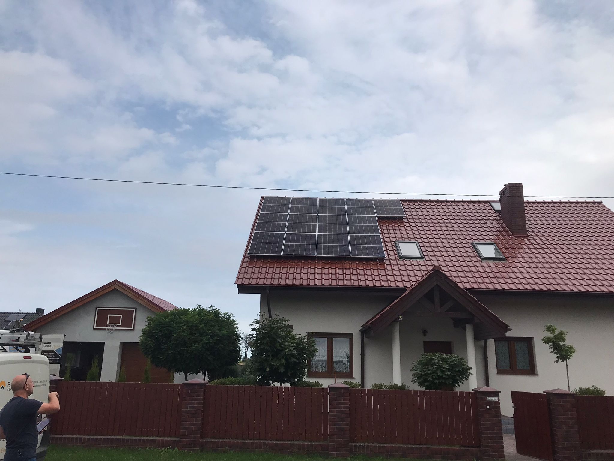 Dom jednorodzinny z czerwoną dachówką i panelami słonecznymi na dachu, widok z poziomu ulicy, z fragmentem zaparkowanego samochodu serwisowego w tle.