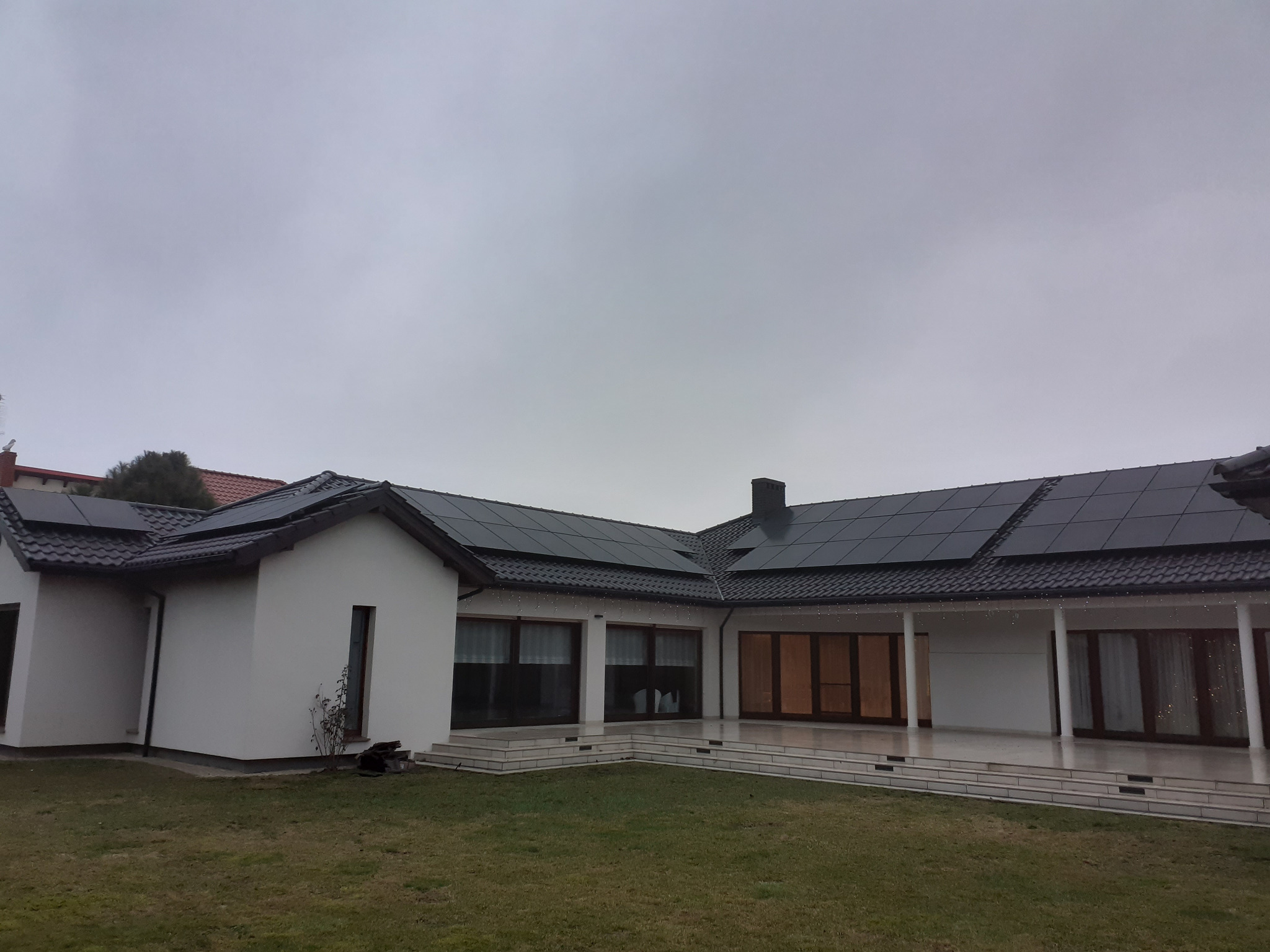 Nowoczesny, biały dom z ciemnymi panelami fotowoltaicznymi na dachu w pochmurny dzień, widok z ogrodu z niską trawą.