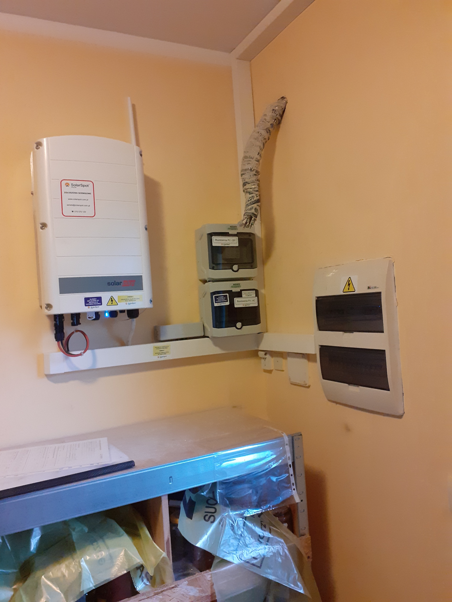Zamontowany inwerter SolarSpot z anteną, obok rozdzielnica elektryczna z żółtym symbolem ostrzegawczym, oraz dodatkowe zabezpieczenia na ścianie o kremowym odcieniu. Przewody zabezpieczone osłoną.