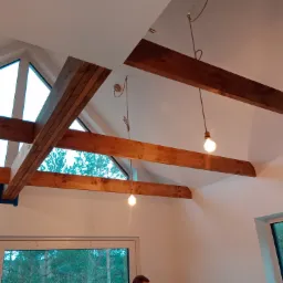 Wnętrze z drewnianymi belkami stropowymi, dużymi oknami z widokiem na las i minimalistycznymi lampami wiszącymi na sznurkach.