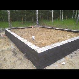 Bs Serwis - Wzniesione mury fundamentowe z pustaków szalunkowych, częściowo pomalowane na czarno, wypełnione piaskiem, na tle lasu. Widoczne elementy instalacji.