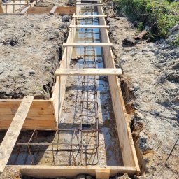 Bs Serwis - Szeroki kadr na wykop z drewnianym szalunkiem i zbrojeniem zalany wodą. Widok z góry na przygotowany wykop pod fundament z elementami zbrojenia.