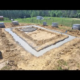 Bs Serwis - Zalane betonem ławy fundamentowe pod budynek na działce z zielenią w tle. Widoczne bloczki betonowe i niebieska rura. Ujęcie z góry, w słoneczny dzień.