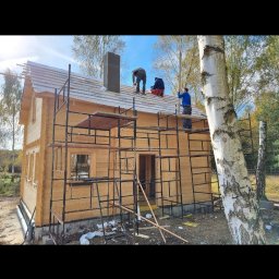 Bs Serwis - Budowa domu z bali: robotnicy na rusztowaniu montują dach. Widoczny komin, drewniana konstrukcja i brzoza na pierwszym planie. Prace w toku, słoneczny dzień.