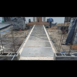 Bs Serwis - Budowa ścieżki z płyt betonowych i żwiru prowadzącej do domu. Widoczne bloczki fundamentowe, zbrojenie i osoba w oddali. Prace w toku, teren budowy.