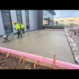 Bs Serwis - Pracownicy w żółtych kamizelkach zacierają świeżo wylany beton na zewnątrz budynku. Widoczne szalunki z różowym styropianem i narzędzia do zacierania betonu.