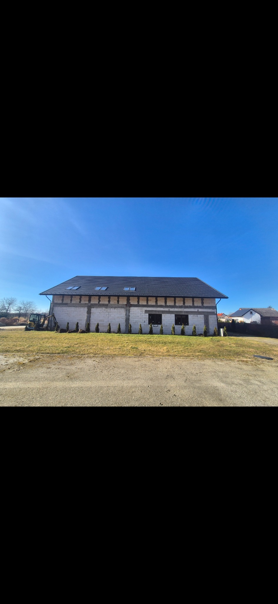 Budynek w trakcie budowy z białych bloczków, z czarnym dachem i małymi oknami dachowymi. Na trawniku posadzone małe tuje. Z boku koparka.