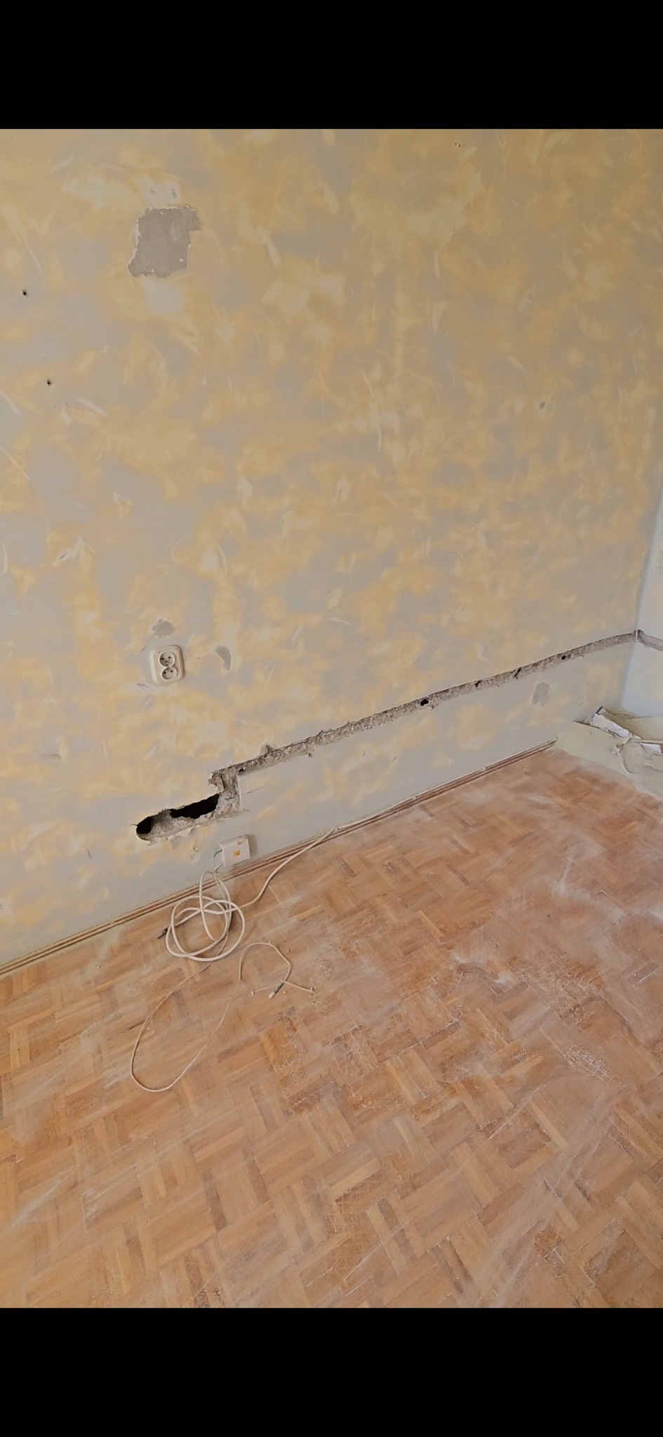 Wnętrze w trakcie remontu: ściana z uszkodzeniami i bruzdą na instalacje elektryczne, widoczny parkiet i kabel. Prace przygotowawcze do położenia instalacji.