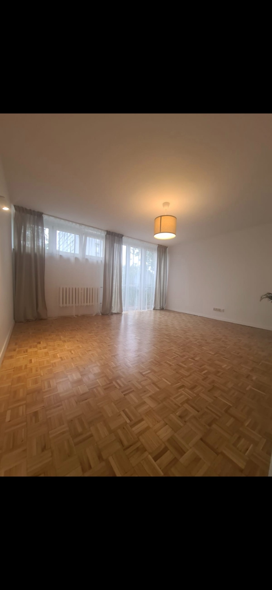 Jasne, przestronne wnętrze z parkietem w jodełkę, zasłonami i lampą. Świeżo wyremontowany pokój z grzejnikiem pod oknem i widocznymi gniazdkami elektrycznymi.