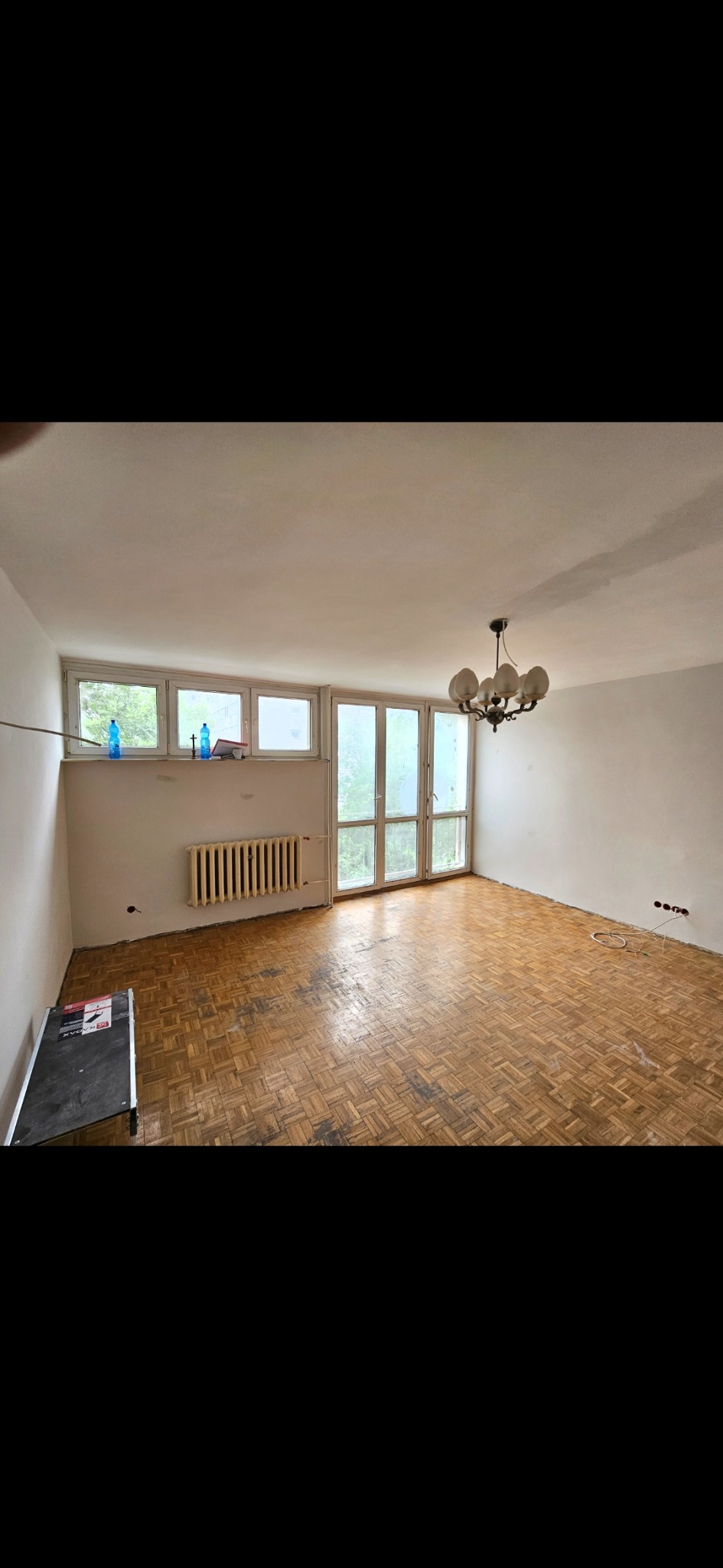 Pusty pokój przed remontem: parkiet w jodełkę z widocznymi śladami użytkowania, grzejnik pod oknem, surowe ściany przygotowane do malowania, wisząca lampa.