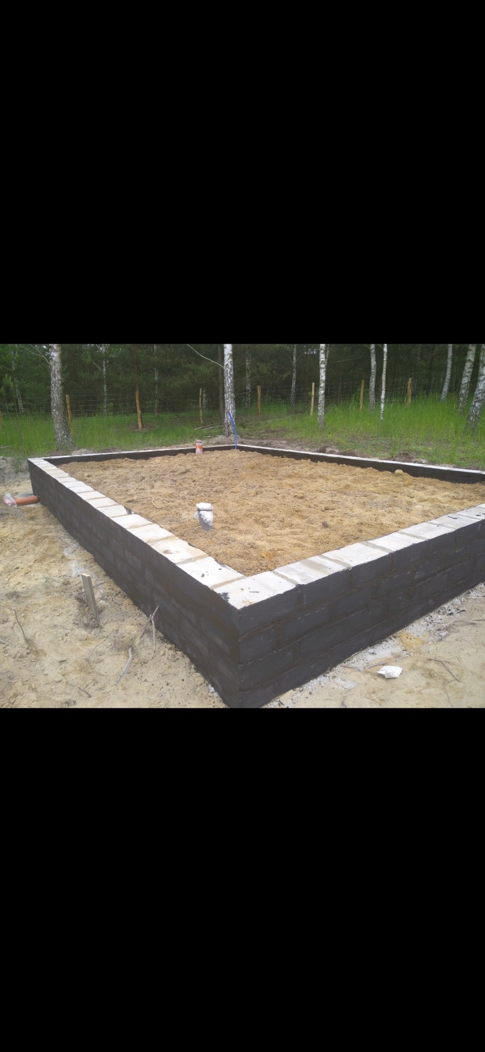 Wzniesione mury fundamentowe z pustaków szalunkowych, częściowo pomalowane na czarno, wypełnione piaskiem, na tle lasu. Widoczne elementy instalacji.