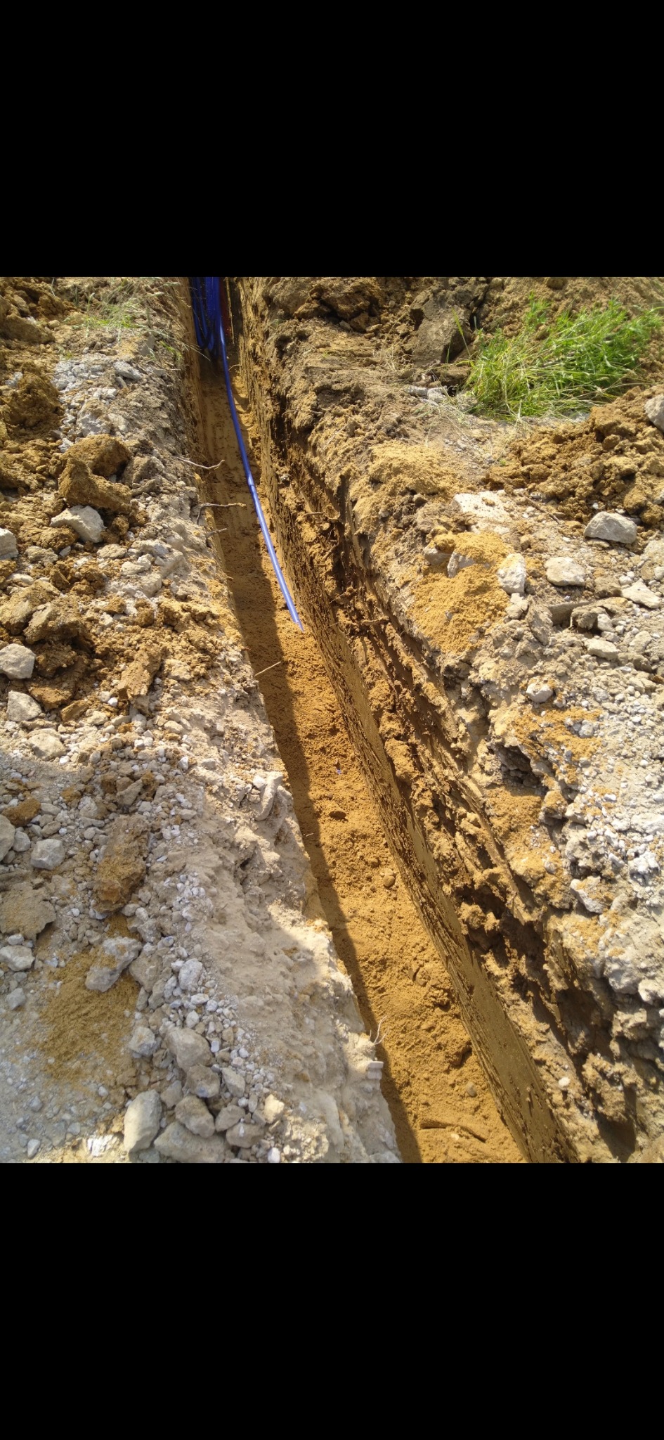 Wąski wykop w ziemi z ułożonymi niebieskimi rurami instalacyjnymi. Ściany wykopu z piasku i kamieni, widoczne ślady po narzędziach. Częściowo widoczna trawa.