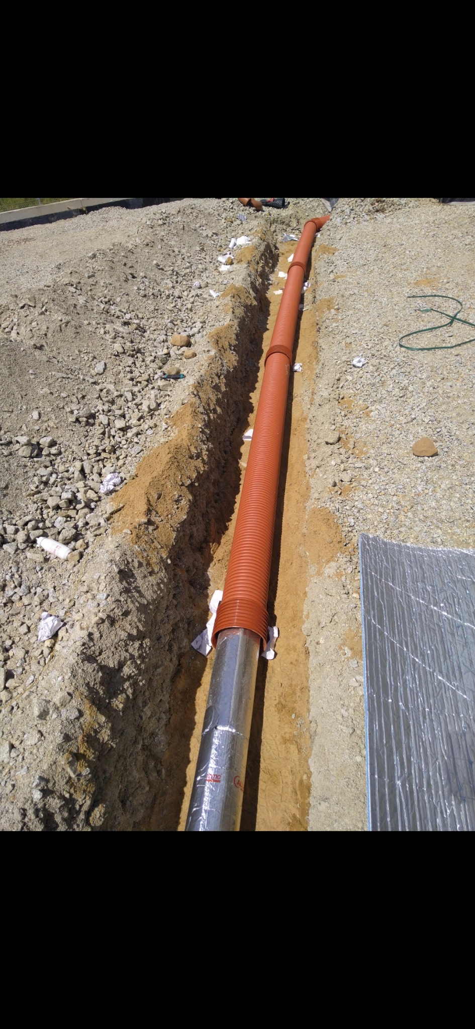 Pomarańczowa rura kanalizacyjna w wykopie, częściowo owinięta folią aluminiową. Widoczne białe podpórki i fragment izolacji. Grunt piaszczysty, kamienisty.