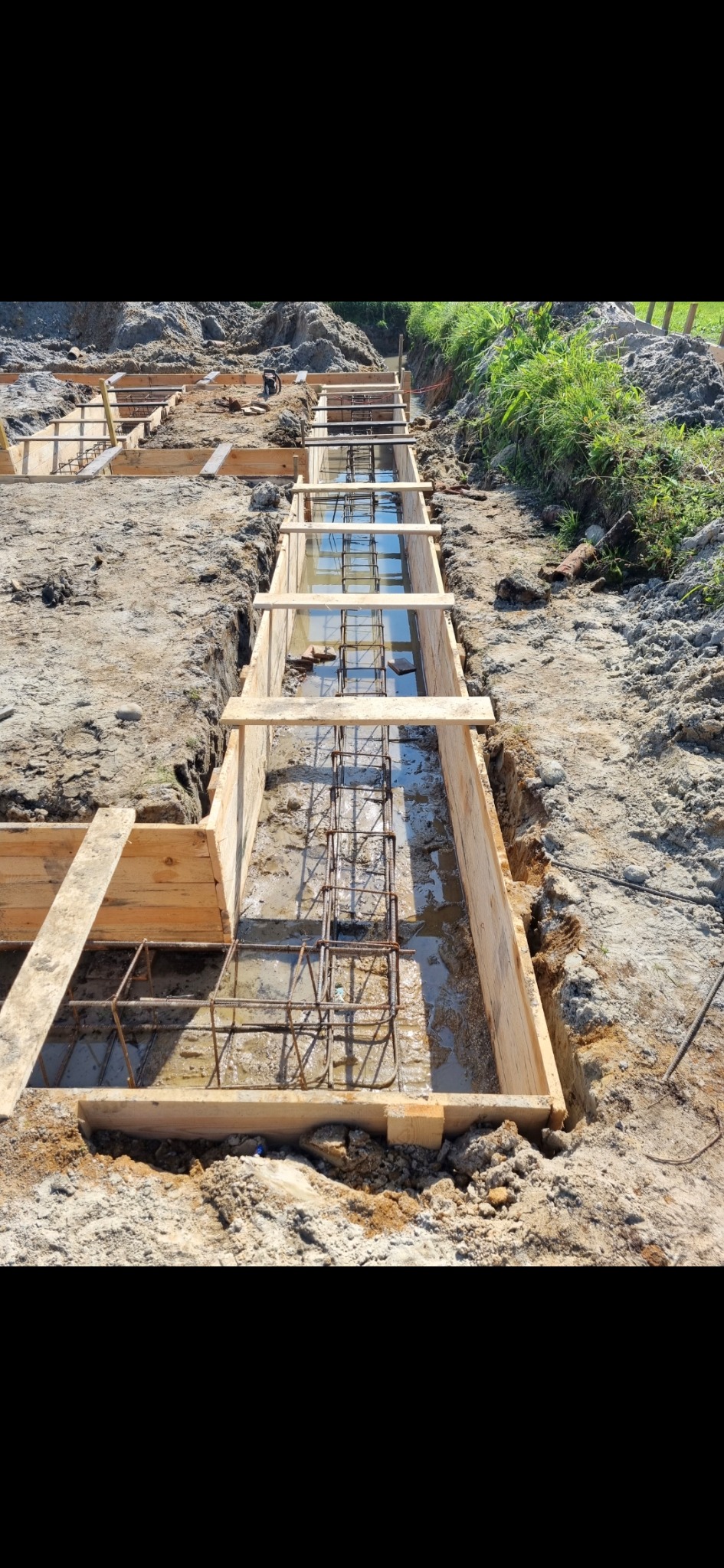 Szeroki kadr na wykop z drewnianym szalunkiem i zbrojeniem zalany wodą. Widok z góry na przygotowany wykop pod fundament z elementami zbrojenia.