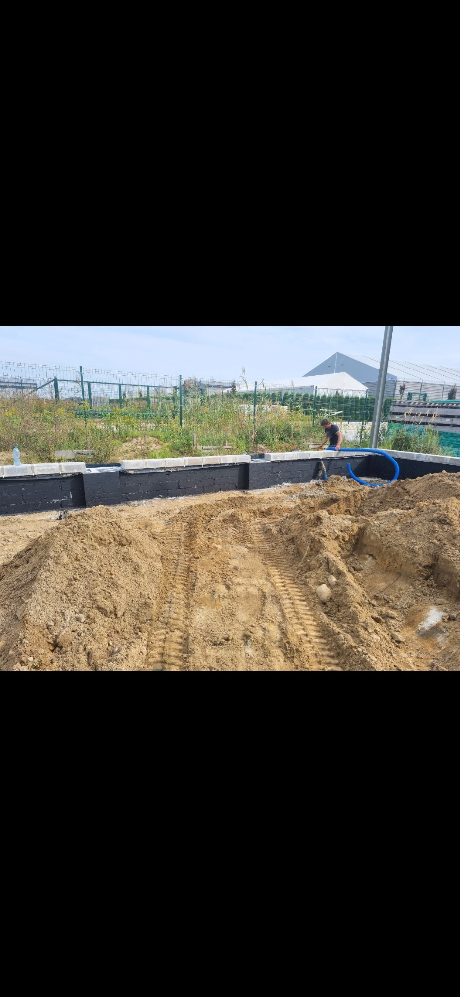 Wykop pod fundamenty z częściowo wzniesionymi ścianami z bloczków, zabezpieczonymi czarną izolacją. Widoczny pracownik i niebieski przewód instalacyjny.