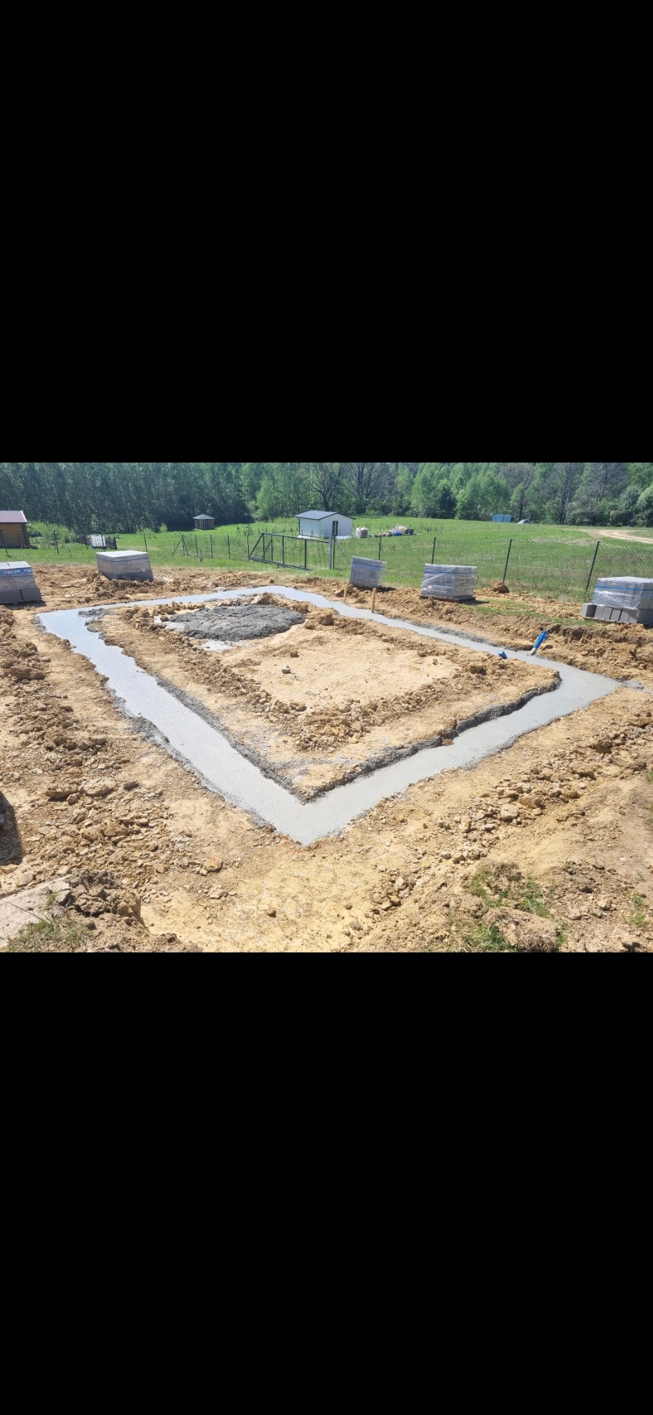 Zalane betonem ławy fundamentowe pod budynek na działce z zielenią w tle. Widoczne bloczki betonowe i niebieska rura. Ujęcie z góry, w słoneczny dzień.