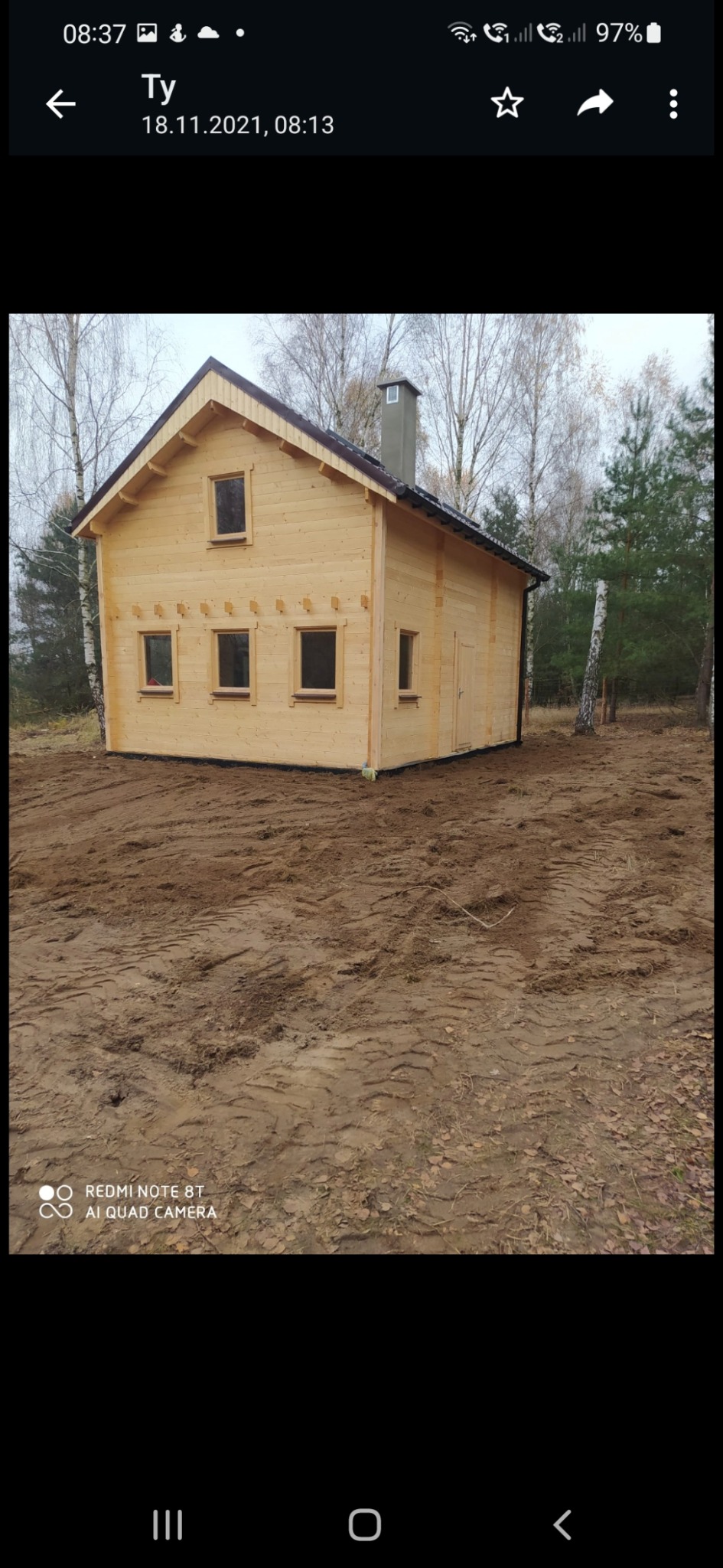 Nowy, drewniany domek letniskowy z kominem, na przygotowanym terenie. Widoczne małe okna i drzwi. Tło stanowią drzewa bez liści. Ujęcie z poziomu gruntu.