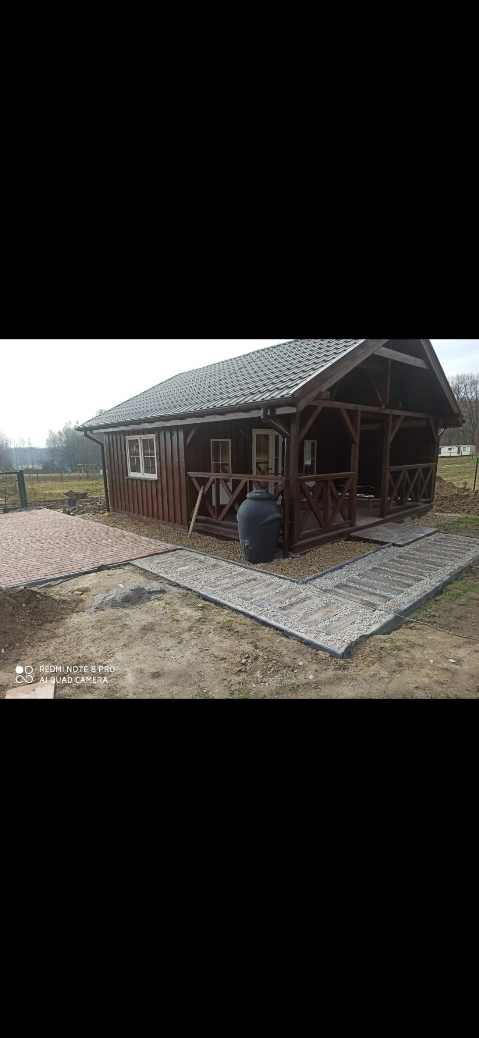 Drewniany domek letniskowy z zadaszonym tarasem, obok ścieżka z płyt i kamieni. Ujęcie z poziomu gruntu, widoczne detale wykończenia i otoczenie.