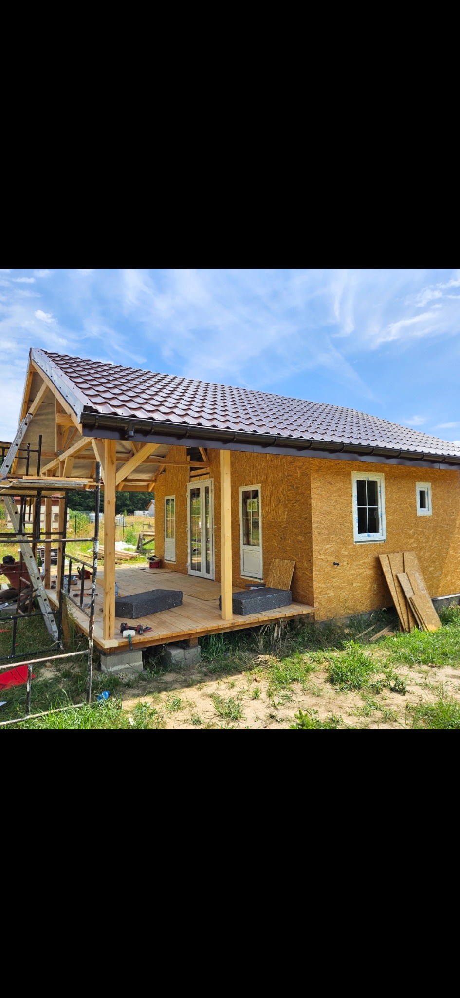 Dom w trakcie budowy z drewnianą konstrukcją, brązowym dachem i ścianami z płyt OSB. Widoczne rusztowania i materiały budowlane. Błękitne niebo w tle.