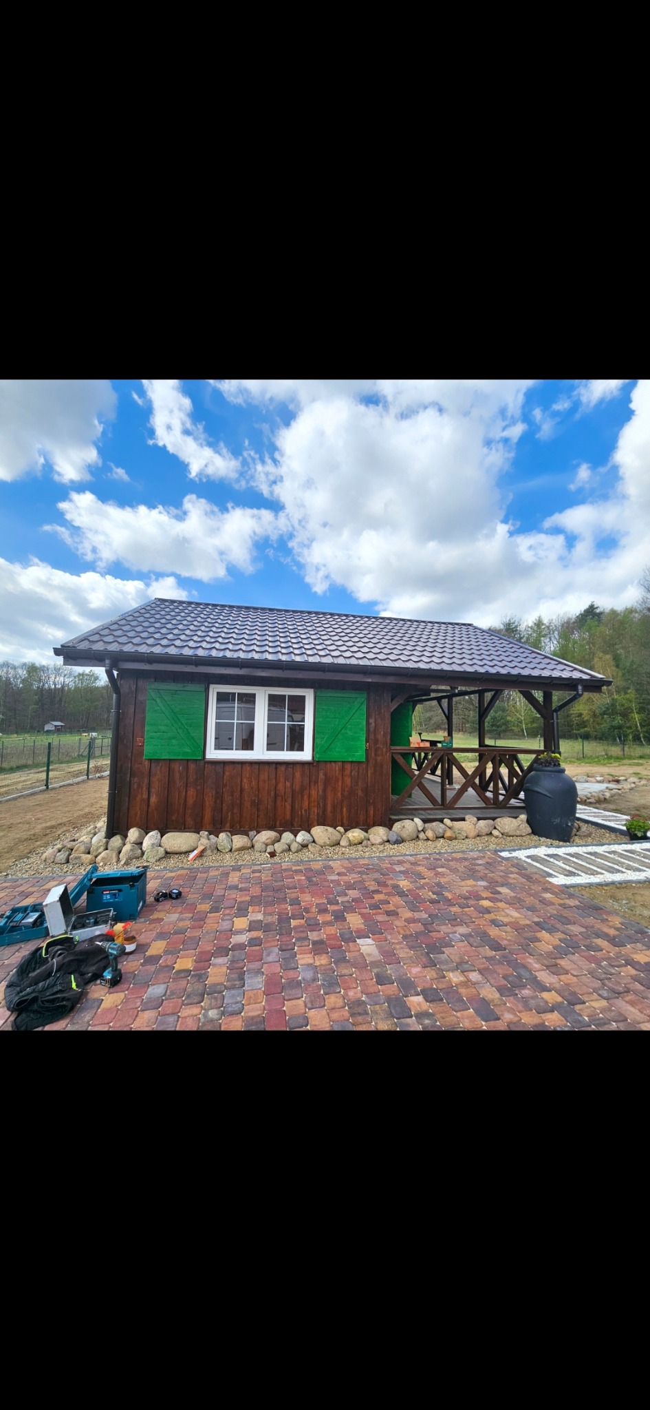 Drewniana altana z zielonymi okiennicami i brązowym dachem, otoczona kostką brukową i kamieniami. Widoczne narzędzia i akcesoria budowlane.