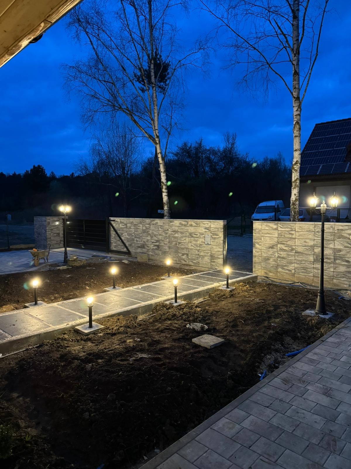 Oświetlona ścieżka z kostki brukowej i lamp ogrodowych, otoczona świeżo przekopaną ziemią, prowadząca do bramy w murze z bloczków betonowych, wieczorne ujęcie.