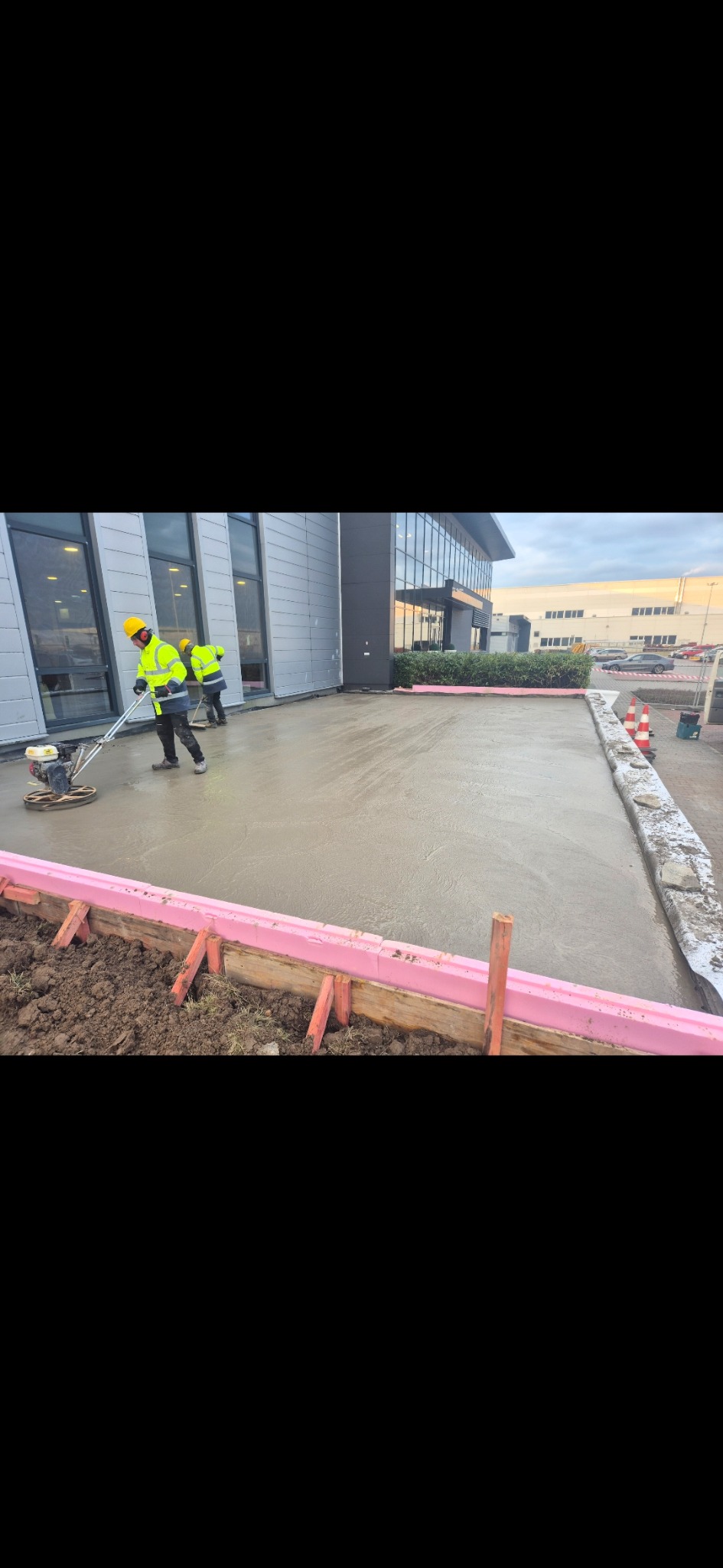 Pracownicy w żółtych kamizelkach zacierają świeżo wylany beton na zewnątrz budynku. Widoczne szalunki z różowym styropianem i narzędzia do zacierania betonu.