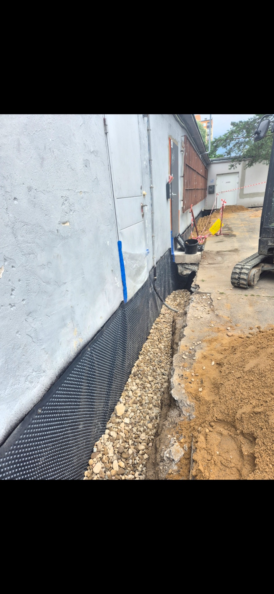 Odkryte fundamenty budynku z izolacją przeciwwilgociową i warstwą drenażową z kamieni. W tle widoczny fragment koparki i elementy zabezpieczenia terenu.