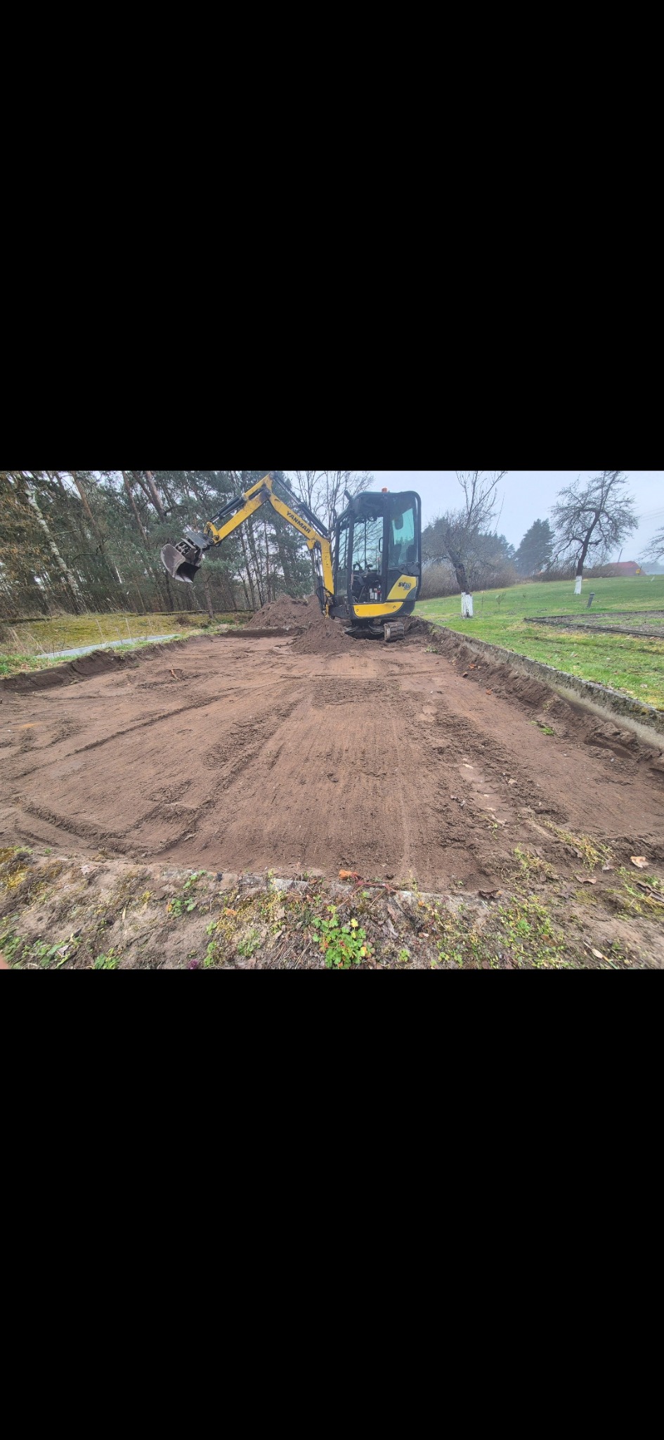 Żółta mini koparka Yanmar na wykopanym, wyrównanym terenie pod budowę. Widoczne ślady opon i wykopane obrzeża. W tle drzewa i trawa.
