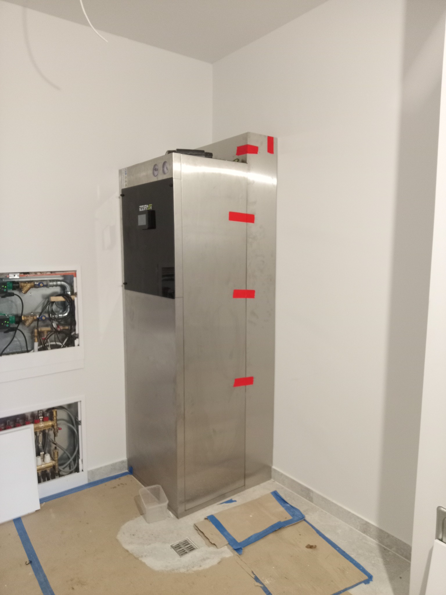 Nowoczesny piec gazowy ze stali nierdzewnej, zamontowany w pomieszczeniu z widoczną instalacją hydrauliczną w ścianie. Widoczne zabezpieczenia podłoża.