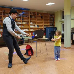 Iluzjonista w czarnym kapeluszu i kamizelce prezentuje trik z liną małej dziewczynce w żółtej koszulce, na tle regałów z książkami.