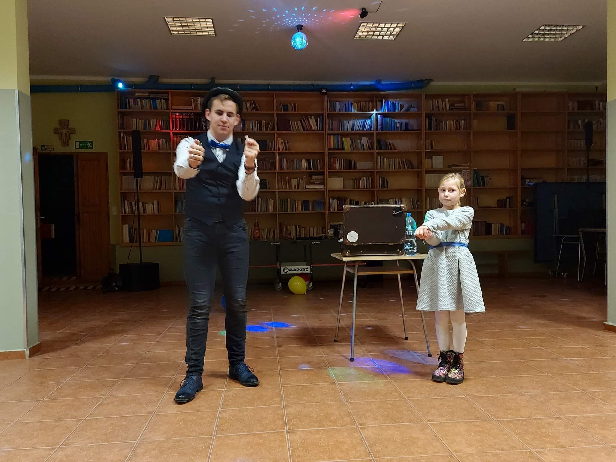 Młody iluzjonista w czarnym meloniku i muszce prezentuje trik przed dziewczynką w sukience, na tle regałów z książkami, oświetlony kolorowymi światłami dyskotekowymi.