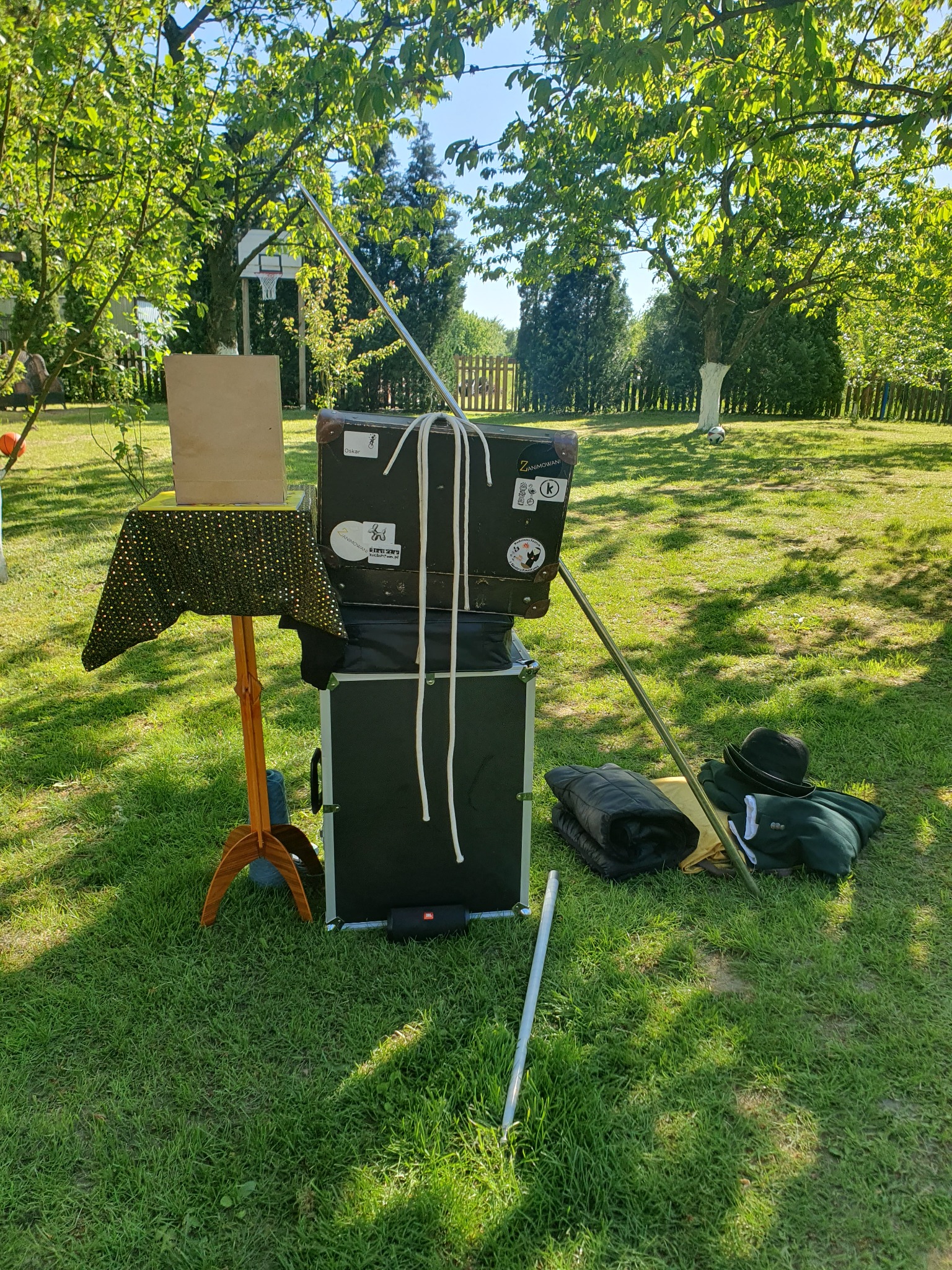 Przygotowania do występu iluzjonisty w plenerze: stół z cekinową narzutą, walizka z naklejkami, rekwizyty, kostium i sprzęt nagłośnieniowy na trawie.