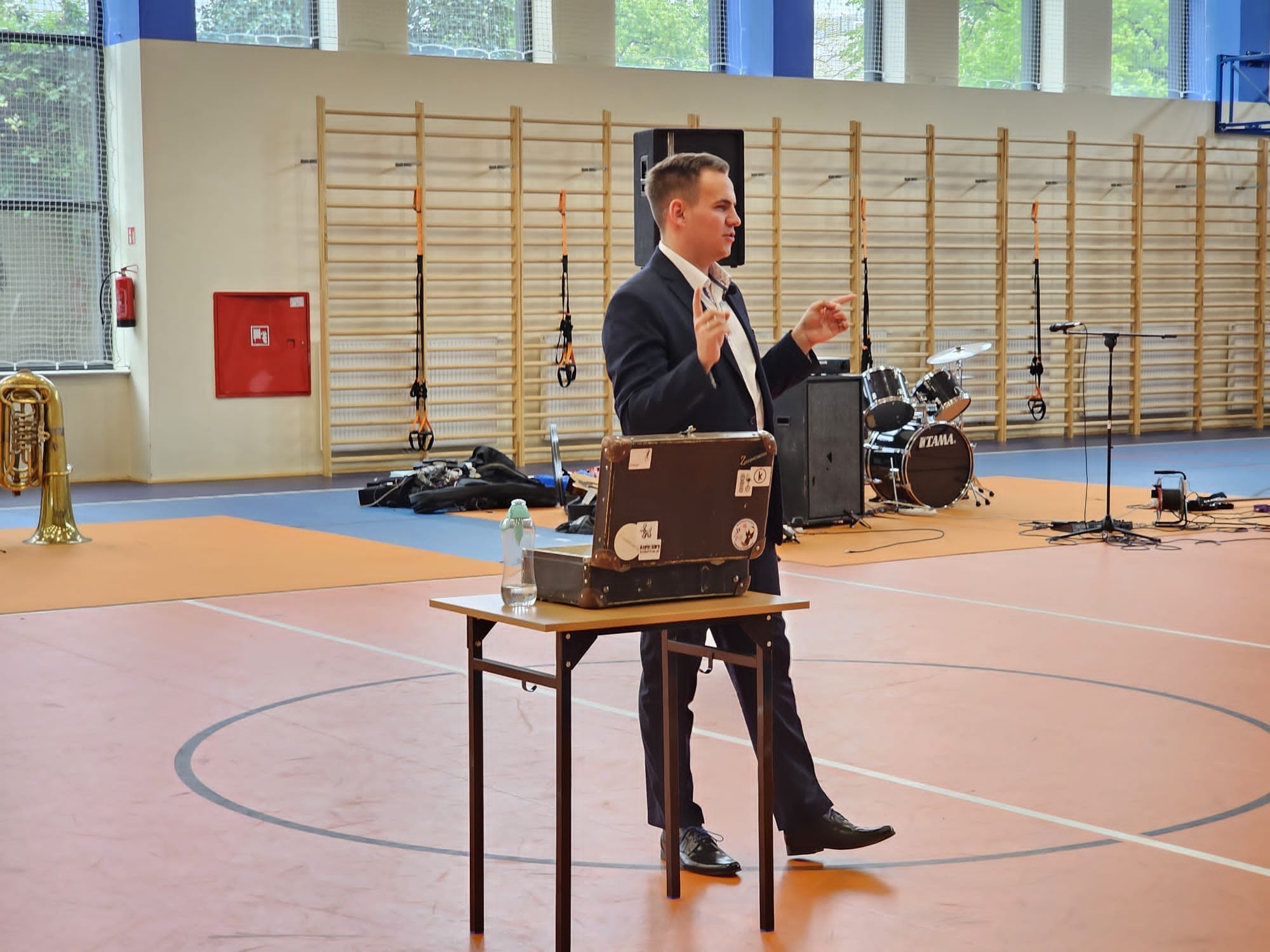 Mężczyzna w garniturze gestykuluje podczas występu na tle sali gimnastycznej z perkusją i trąbką, kuferki na stole.