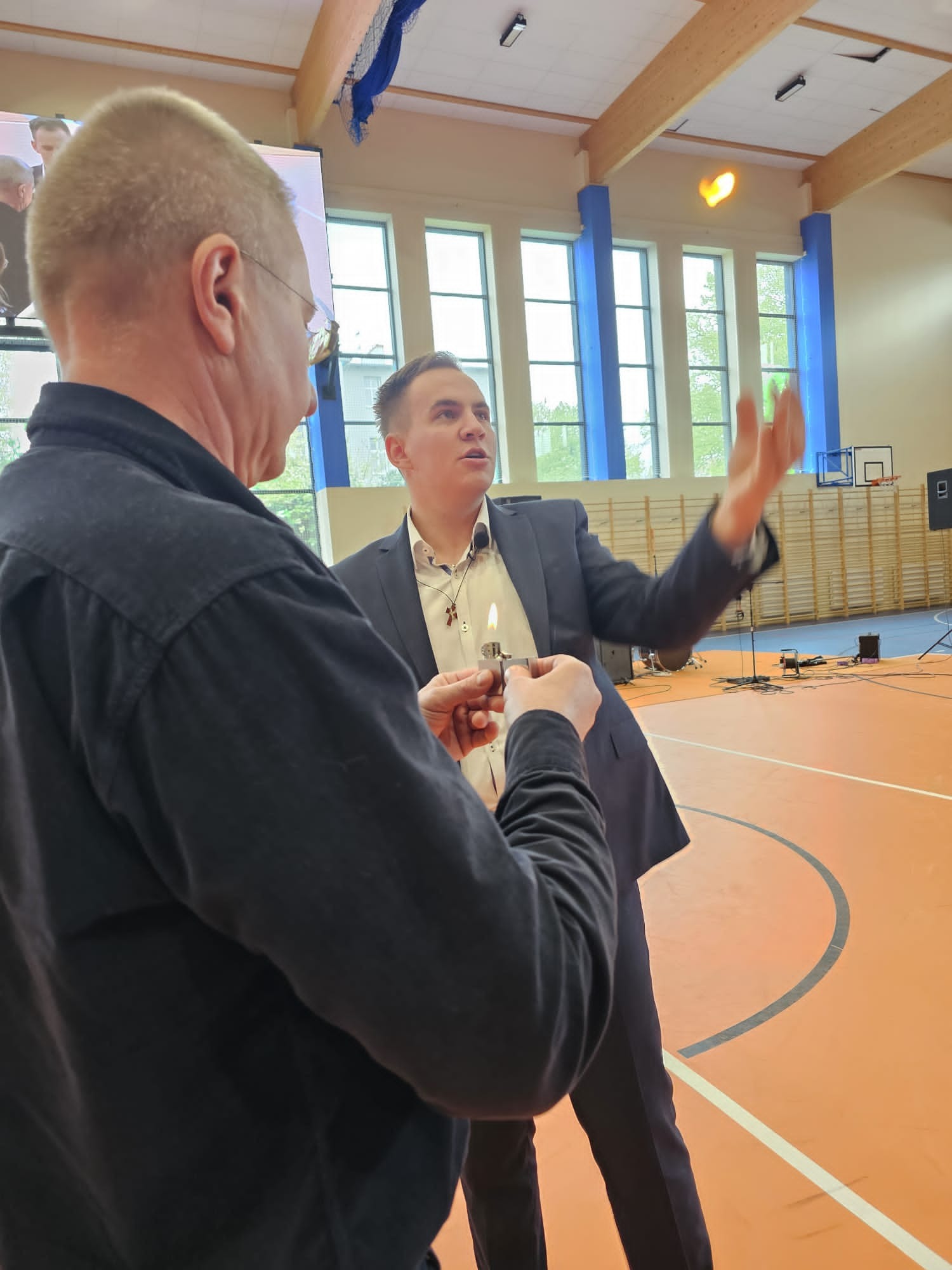 Mężczyzna w garniturze wykonuje trik z ogniem, zapalniczka w dłoni, płomień unosi się w powietrzu, w tle sala gimnastyczna.