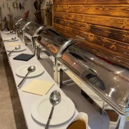 Ustawiony stół szwedzki z podgrzewaczami, talerzami, łyżkami i sosjerkami z sosem, w tle drewniana ściana i dekoracje marynistyczne.