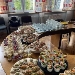 St&oacute;ł okrągły zastawiony r&oacute;żnorodnymi przekąskami: mini kanapeczki z łososiem i figami, sałatki w kubeczkach, desery z owocami, tortille, oraz stosy białych filiżanek z logiem Costa Coffee, widok...