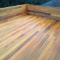 Wykończony taras z desek o zróżnicowanej kolorystyce drewna, z niską ścianką w tle, otoczony zielenią drzew.