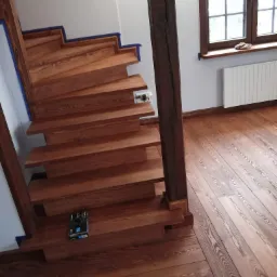 Drewniane schody i podłoga po cyklinowaniu, zabezpieczone niebieską taśmą malarską na krawędziach, z widoczną puszką produktu do obróbki drewna na jednym ze stopni.