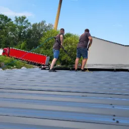 Dwóch mężczyzn czyści dach hali przemysłowej za pomocą kijów teleskopowych z podłączonymi wężami, w tle zaparkowana czerwona ciężarówka i zieleń.