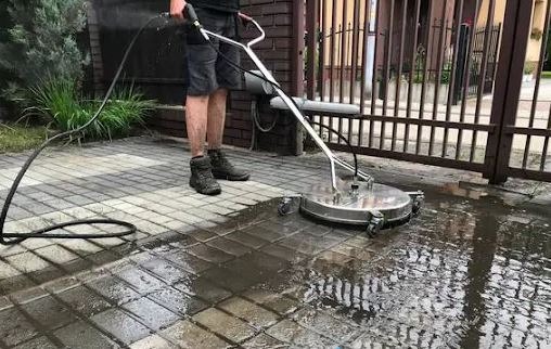 Mężczyzna w roboczych butach czyści kostkę brukową za pomocą okrągłej myjki ciśnieniowej; widoczny efekt czyszczenia na mokrej powierzchni.