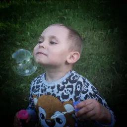 Chłopiec w bluzie z jelonkiem dmucha bańki mydlane na tle zielonej trawy.