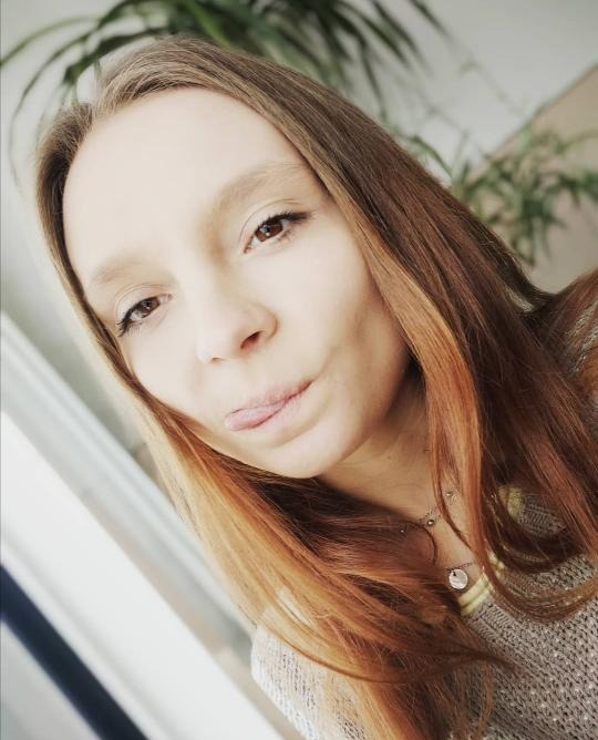 Portret młodej kobiety z wyciągniętym językiem, z długimi, brązowo-blond włosami i delikatnym makijażem, na tle okna i zielonej rośliny.