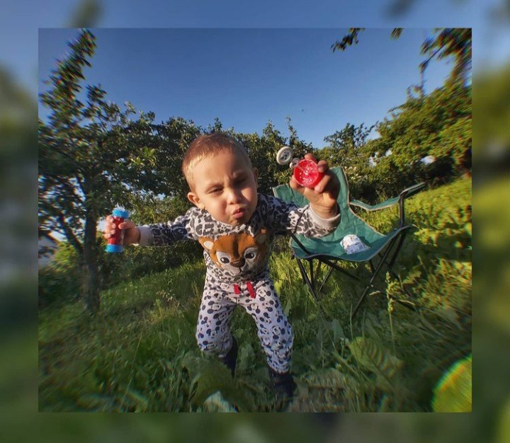 Mały chłopiec w piżamie z motywem zwierzęcym trzyma w dłoniach zabawki do robienia baniek mydlanych na tle zielonego ogrodu i składanego krzesła turystycznego.