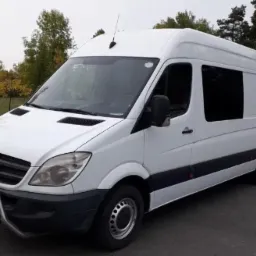 Biały, wysoki furgon Mercedes-Benz Sprinter z chromowanym orurowaniem przedniego zderzaka, zaparkowany na asfalcie w otoczeniu drzew.