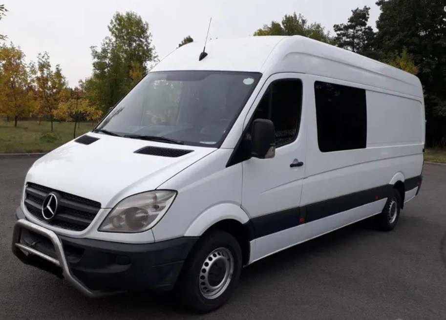 Biały, wysoki furgon Mercedes-Benz Sprinter z chromowanym orurowaniem przedniego zderzaka, zaparkowany na asfalcie w otoczeniu drzew.