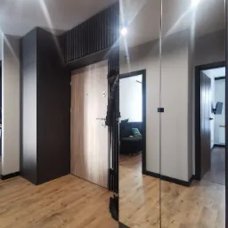 Nowoczesny przedpokój z drewnianą podłogą, lustrzaną szafą wnękową, grafitową zabudową, drewnianymi drzwiami wejściowymi i czarnym wieszakiem z kurtką.