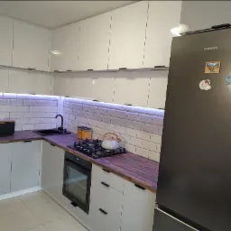 Nowoczesna kuchnia z białymi szafkami, ciemnym blatem, podświetleniem LED pod szafkami wiszącymi oraz czarnym zlewem i piekarnikiem, uzupełniona o lod&oacute;wkę Samsung z magnesami.