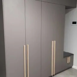 Szara zabudowa meblowa z drewnianymi uchwytami, obejmująca szafy i siedzisko, na tle szarych płytek podłogowych oraz białej ściany z panelem sterowania.