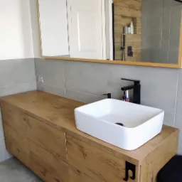 Nowoczesna łazienka z drewnianą szafką pod umywalką i prostokątną białą umywalką nablatową, czarna bateria, lustro z drewnianą ramą odbijające prysznic z drewnianymi panelami.