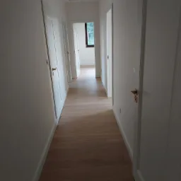 Długi korytarz z jasnym parkietem ułożonym w deski, białymi ścianami i białymi drzwiami z minimalistycznymi, drewnianymi klamkami, widok w głąb z oknem na końcu.